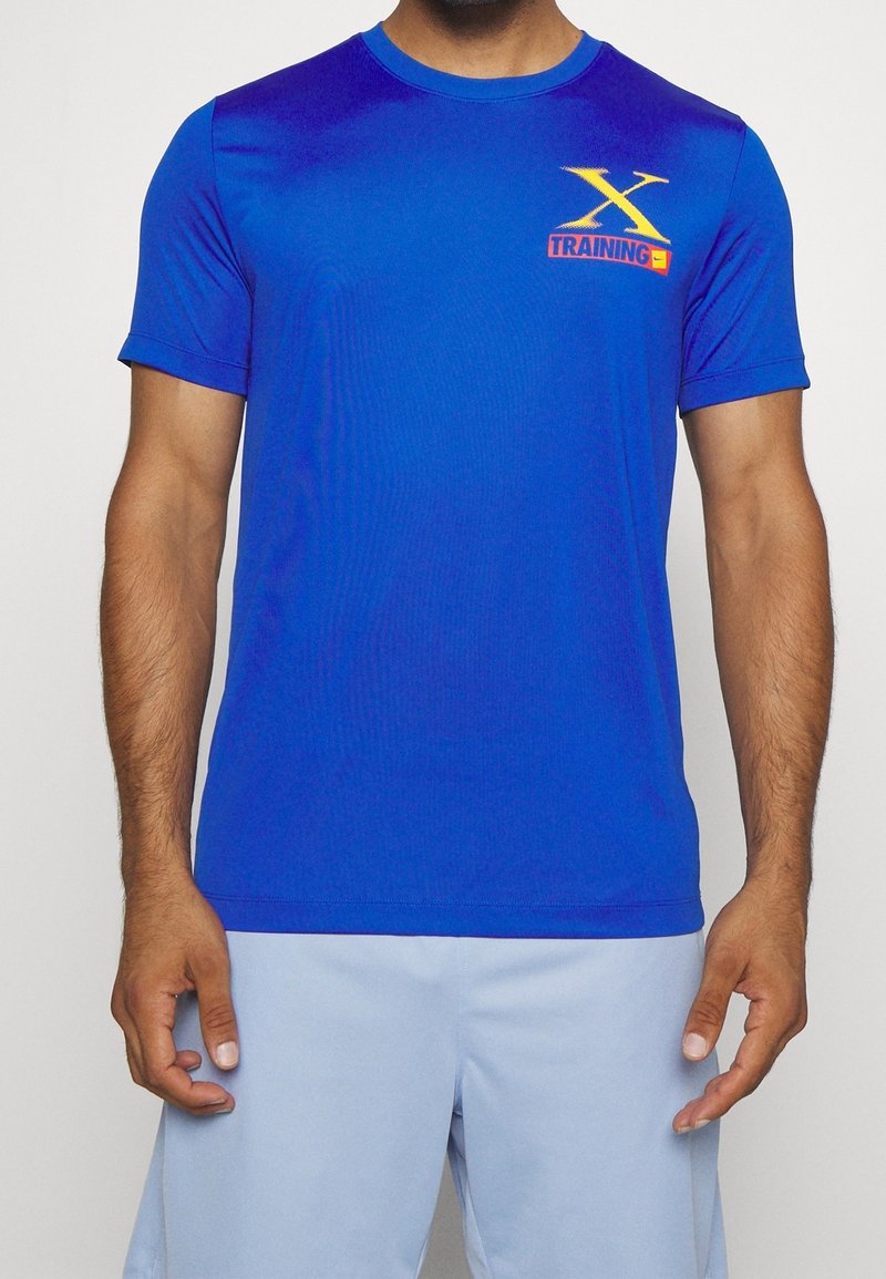 T-shirt de sport bleu à col rond, manches courtes, avec un logo imprimé "TRAINING" en jaune et orange sur le côté gauche de la poitrine. Texture lisse.