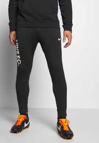 Svarta träningsbyxor med slim fit, med en vit Nike-logotyp och texten "Nike F.C." på sidan. Bärs tillsammans med svarta och orange fotbollsskor.