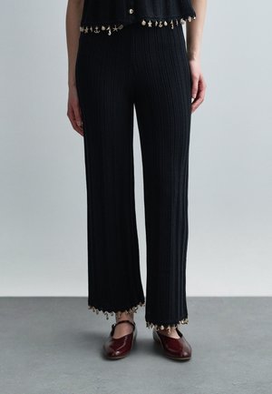 Žena nosí čierne rebrované nohavice s širokými nohavicami, ktoré majú malé zlaté ozdoby na lemovaní, a bordové topánky Mary Jane na sivom podlahe.