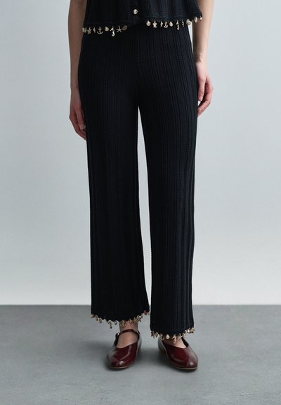 Femme portant un pantalon large côtelé noir avec de petits charms dorés aux ourlets et des chaussures Mary Jane bordeaux sur un sol gris.
