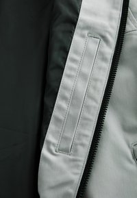 Furnizată cu o căptușeală de culoare verde închis, cu un exterior texturat de culoare gri. Dispune de un fermoar și o pungă rectangulară pe partea stângă.