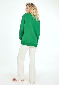 Žalias per didelis džemperis su apvalia iškirpte ir gumbuotomis rankovėmis, derinamas su baltomis klostuotomis kelnėmis, matomas iš nugaros.