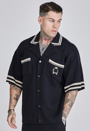 Camisa de manga corta en azul marino con rayas blancas en el cuello y las mangas, tejido texturizado, dos bolsillos en el pecho y un detalle de logo en el bolsillo izquierdo.