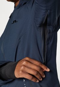 Chaqueta deportiva azul marino hecha de tela ligera, con cremallera lateral, textura suave y detalle de logo cerca del hombro.