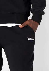 Schwarzer Sweatshirt und Jogginghose mit gerippten Bündchen, seitlichen Taschen und einem weißen Logo mit der Aufschrift "SIXTH JUNE" in Blau auf der Hose.