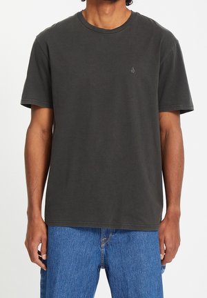 Homme portant un t-shirt uni gris foncé à manches courtes et un jean bleu, debout devant un fond blanc.