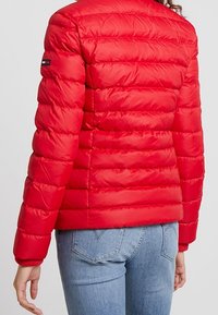 Rød quilted jakke med et lille logo på ærmet, båret af en person med lyseblå jeans, vist fra ryggen og op til taljen.