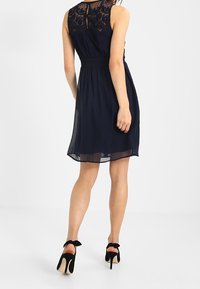 Marinblå klänning med en genomskinlig spets rygg, åtsittande midja och svävande kjol. Bärs med svarta högklackade skor som har rosettaccent.