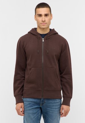 Brauner Kapuzenpullover mit Reißverschluss aus weichem Stoff, mit einer Fronttasche, Kordelzugkapuze und gerippten Bündchen. Wird über einem dunklen Shirt getragen und mit blauer Jeans kombiniert.