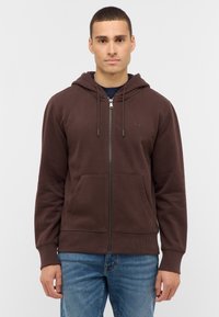 Brauner Kapuzenpullover mit Reißverschluss aus weichem Stoff, mit einer Fronttasche, Kordelzugkapuze und gerippten Bündchen. Wird über einem dunklen Shirt getragen und mit blauer Jeans kombiniert.