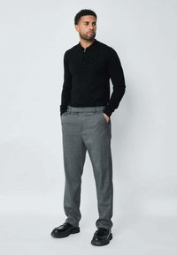 Haut noir à manches longues avec un col zippé, associé à un pantalon à carreaux gris clair et des chaussures noires. La tenue présente une coupe décontractée et une texture subtile.