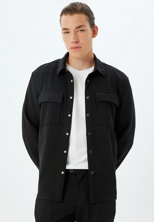 Zwarte button-up shirtjas met twee borstzakken, drukknoopsluitingen en een gladde structuur, gedragen over een wit T-shirt.