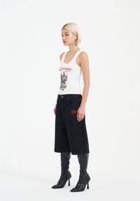 Valkoinen tank top, jossa on graafinen printti ja punaiset tekstit, yhdistettynä mustiin leveisiin housuihin, joissa on punaista brodeerausta, sekä korkeisiin mustiin stiletto-kenkiin.