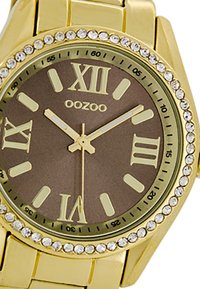 Montre-bracelet dorée avec un cadran marron, index en chiffres romains et accents en cristal autour de la lunette. Équipée d'un bracelet métallique et de aiguilles lumineuses.