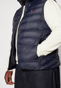 Man som bär en marinblå kviltad väst med röd logotyp över en vit långärmad skjorta, mörka jeans, med en hand i västfickan.