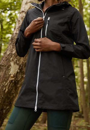 Person, der eine schwarze Jacke mit weißem Reißverschluss trägt und diese im Freien inmitten eines grünen Waldes zumacht.