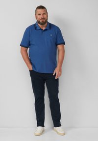 Blauwe polo met kraag en marineblauwe afwerking, gedragen met donkere jeans en witte sneakers. De stof lijkt glad en casual.