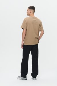 Junger Mann steht, mit dem Rücken zugewandt, trägt ein braunes T-Shirt mit kurzen Ärmeln, schwarze Hosen und grau-weiße Sneakers vor einem schlichten weißen Hintergrund.