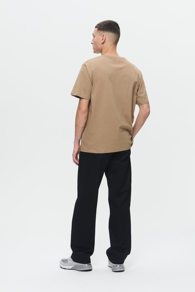 Ung mand står, vender væk, iført en brun kortærmet t-shirt, sorte bukser og grå og hvide sneakers på en ensfarvet hvid baggrund.
