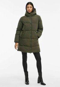 Piumino verde oliva con zip e cappuccio. Caratterizzato da ampie sezioni trapuntate e tasche laterali. Abbinato a jeans skinny neri e stivali con tacco.