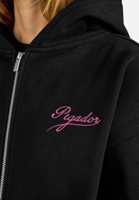 Felpa nera con zip in cotone, caratterizzata da un logo ricamato in rosa "Pegador" sul lato sinistro del petto. Ha una finitura testurizzata e un grande cappuccio.