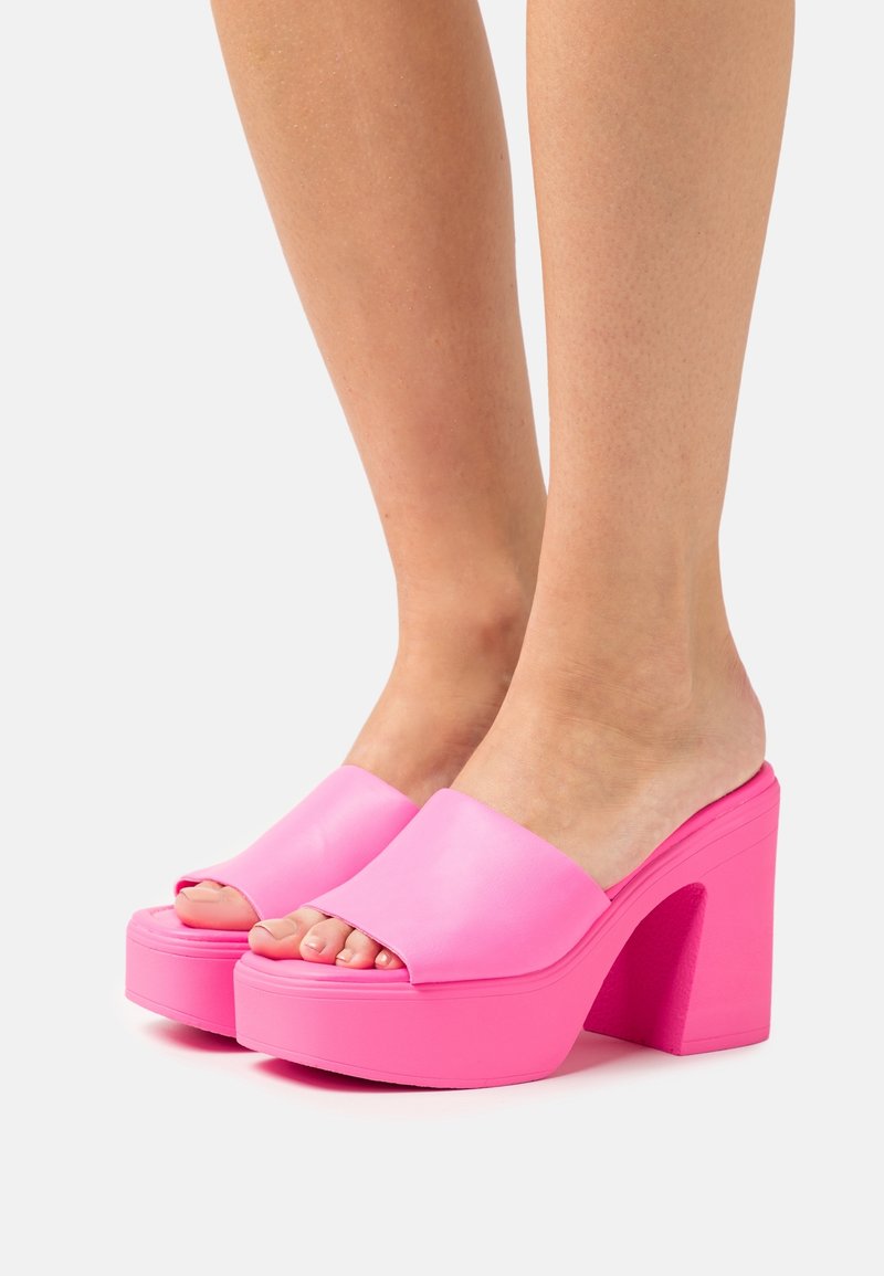 Personne portant des sandales à plateforme à talons hauts ouvertes à l’avant de couleur rose vif, pieds nus, sur fond blanc.