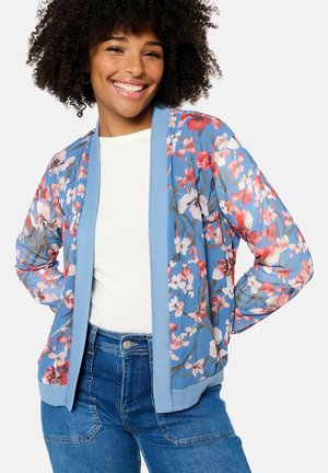 Cardigan léger à motif floral avec une base bleue douce, design à ouverture frontale, poignets et ourlet côtelés, associé à un haut blanc.