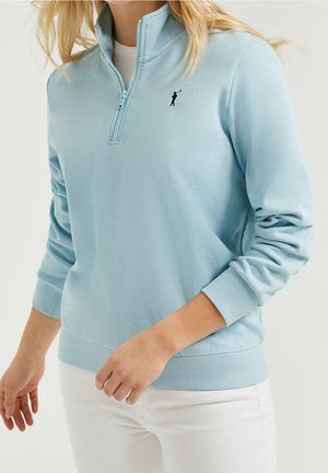 Mujer que lleva un jersey azul claro con cremallera hasta un cuarto, mangas largas y pantalones blancos, con un pequeño logotipo negro de golfista en el pecho.