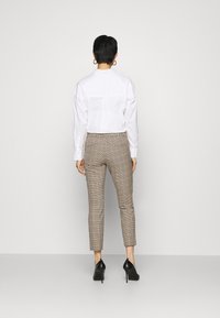 Vit skjorta med lång ärm, kombinerad med bruna rutiga slim-fit byxor och svarta högklackade skor. Bakifrån, enkel bakgrund.