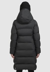 Piumino nero con cappuccio, caratterizzato da sezioni trapuntate. Il materiale appare liscio e lucido, con una silhouette lunga e cerniere laterali.