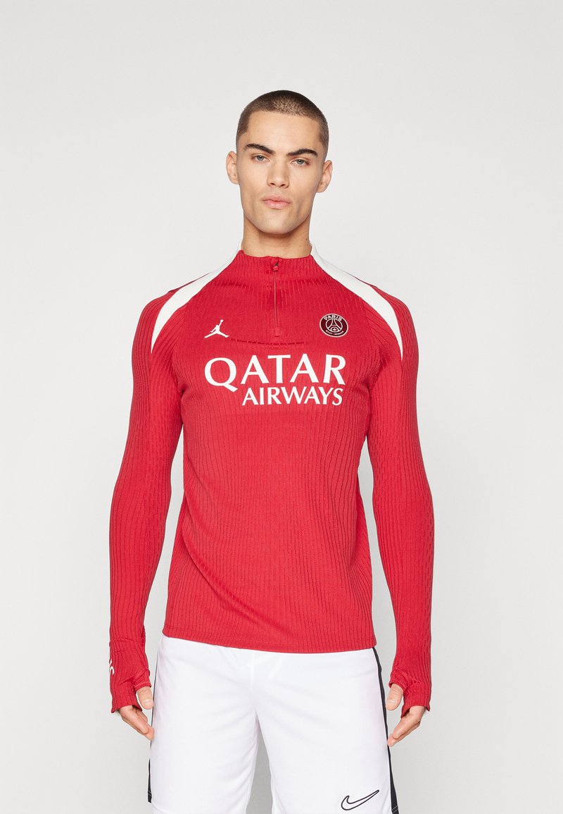 Piros, hosszú ujjú sport póló bordázott textúrával, magas nyakkivágással és hüvelykujj lyukakkal. Fehér díszítésekkel és logóval rendelkezik a mellkason.