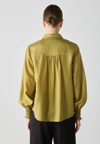 Blusa a maniche lunghe verde oliva con finitura testurizzata, dotata di colletto e polsini elasticizzati. Vestibilità ampia con dettaglio arricciato sul retro.