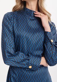 Bluzka w niebiesko-czarny wzór w diamenty, z wysokim dekoltem, długimi rękawami i złotymi guzikami na mankietach. Gładki, błyszczący materiał.