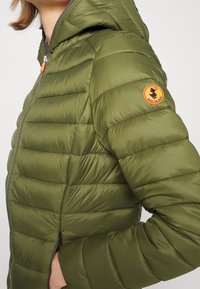 Oliven grøn, let pufferjakke med hætte, der har vandret quiltning, lynlås foran og et orange cirkulært logo på ærmet.