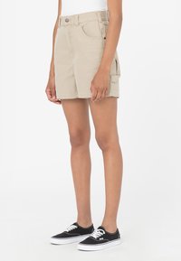 Short en jean beige taille haute avec poche latérale, porté avec des baskets basses noires sur une personne debout.