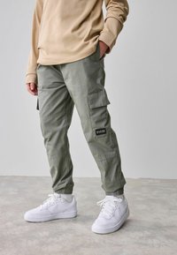 Groene cargo broek met elastische manchetten, zijzakken, een zwart etiket, gedragen met witte Nike schoenen op een lichtgrijze vloer.