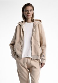 Hoodie beige en matériau doux avec poches avant, cordon de serrage ajustable et bords côtelés. Porté sur un t-shirt blanc.