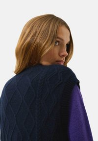 Granatowy, bezrękawnik w sploty kablowe z teksturowanymi wzorami w romby, noszony na purpurowej bluzce z długim rękawem. Brzeg i dekolt wykończone prążkowanym splotem.