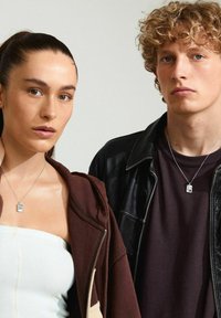 To personer står tæt sammen og bærer sølvvedhæng. Kvinden har en brun zip-up hoodie på over en hvid top; manden har en sort læderjakke på over en mørk skjorte.