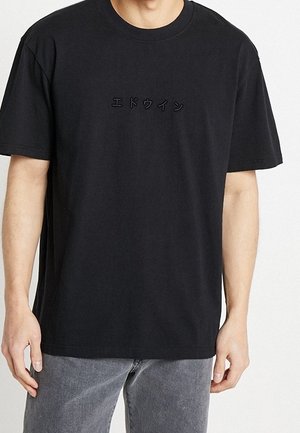 Czarny t-shirt z krótkim rękawem z małym czarnym japońskim napisem wyhaftowanym na piersi, noszony przez osobę w szarych dżinsach na jednolitym tle.