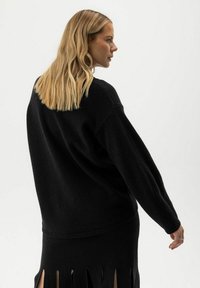 Blonde Frau in einem schwarzen oversized Pullover und passendem Rock mit Schlitz dreht sich seitlich vor einem schlichten hellen Hintergrund.