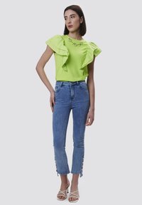 Top arricciato verde lime con scollatura decorativa, abbinato a jeans skinny azzurri chiari con dettagli a bottone all'orlo e sandali bianchi con tacco a blocco.