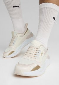 Sneakers PUMA w kolorze beżowym i kremowym z siateczkowymi i skórzanymi panelami. Okrągły czubek, wyściełana podeszwa i paski akcentujące. Noszone z białymi skarpetkami w prążki.