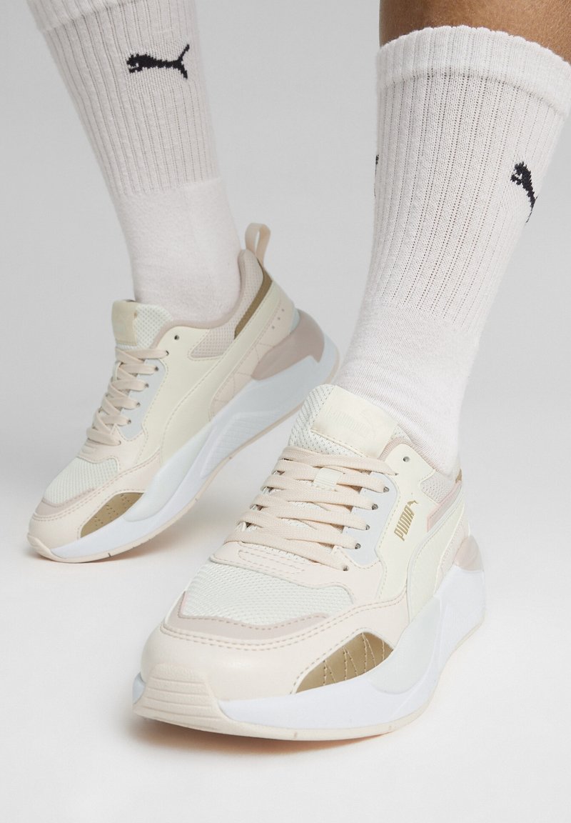 Sneakers PUMA w kolorze beżowym i kremowym z siateczkowymi i skórzanymi panelami. Okrągły czubek, wyściełana podeszwa i paski akcentujące. Noszone z białymi skarpetkami w prążki.