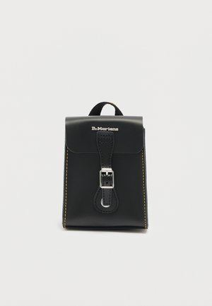Sac à dos Dr. Martens en cuir noir avec fermeture à boucle argentée, coutures jaunes le long des côtés et poignée supérieure, sur fond blanc uni.