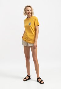 T-shirt in cotone gialla con un design grafico circolare sul petto, abbinata a pantaloni beige e sandali neri. Outfit semplice e casual.