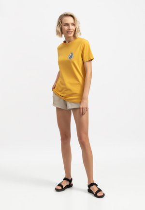 T-shirt in cotone gialla con un design grafico circolare sul petto, abbinata a pantaloni beige e sandali neri. Outfit semplice e casual.