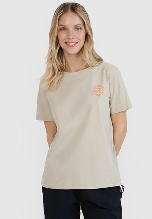 Uśmiechnięta kobieta z długimi blond włosami, ubrana w beżowy t-shirt z pomarańczowym napisem "EVERYDAY" na piersi i czarne spodnie na białym tle.