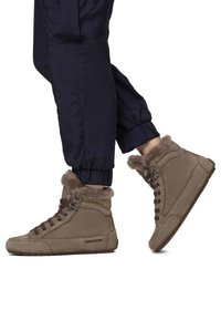 Sneaker alte in pelle marrone con colletto in pelliccia sintetica, lacci marroni e suola in gomma piatta. Indossati con pantaloni jogger blu.