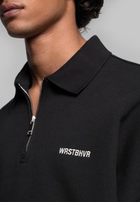 Polo noir à fermeture éclair avec col, présentant une fermeture éclair argentée et le texte brodé en blanc "WRSTBHVN" sur le côté gauche.